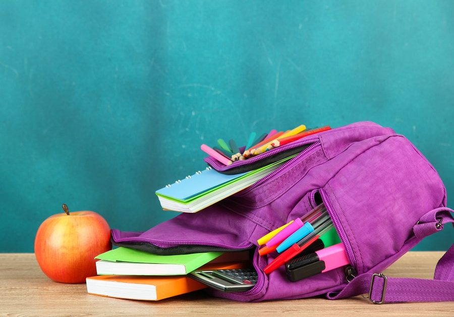 Purple backpack with school supplies on wooden table on green desk background