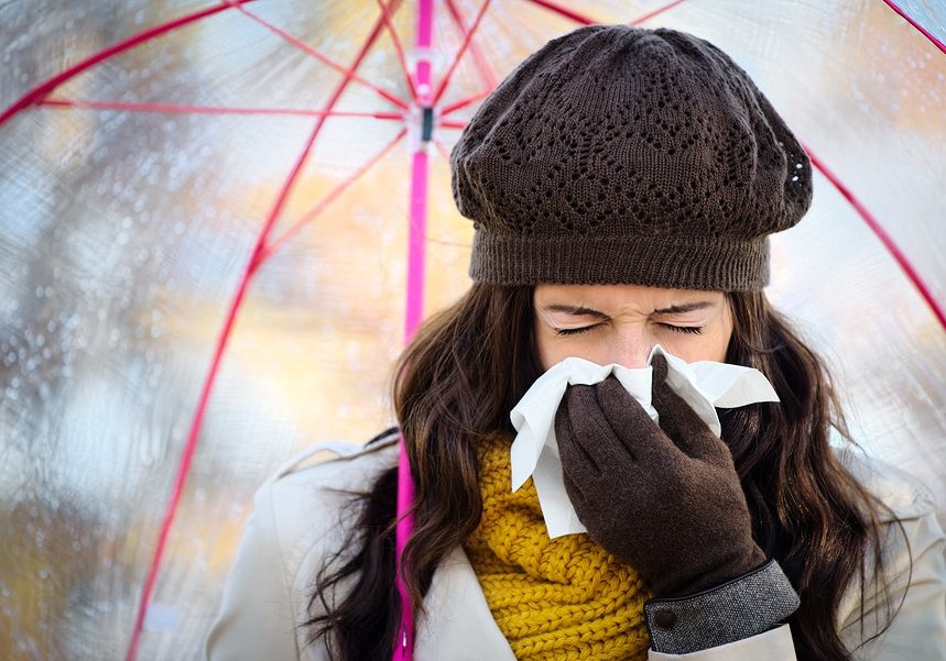 Woman with cold or flu coughing and blowing her nose with a tissue under autumn rain. Brunette female sneezing and wearing warm clothes.