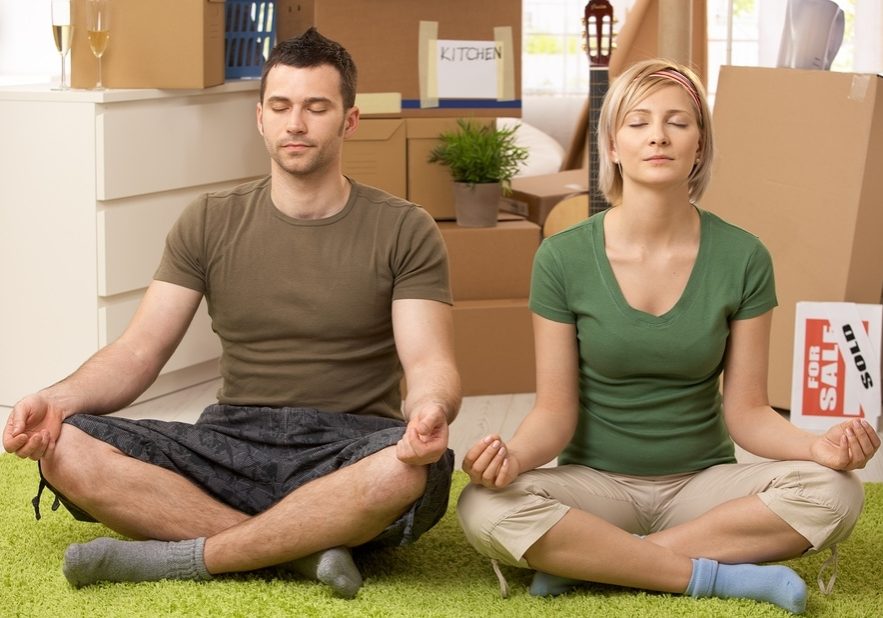 Young couple doing yoga meditation in new house after moving, sitting in middle of boxes.