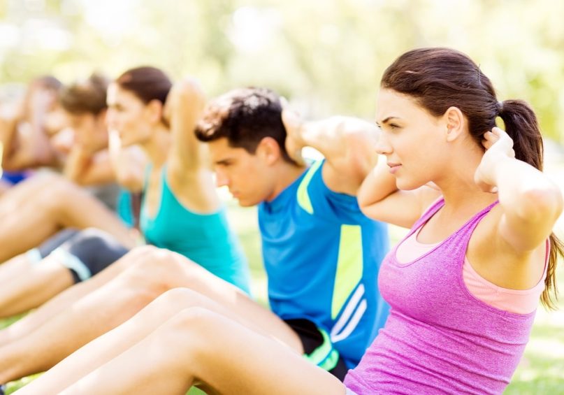 Beautiful young woman with friends doing sit-ups in park. Horizontal shot.